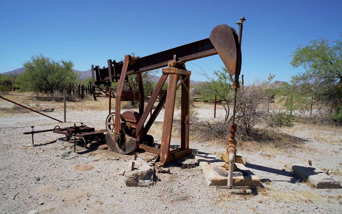 Rusty oil pump jack standing in a dry, scrub-covered landscape, with mountains in the distance beneath a clear blue sky.