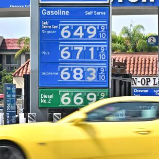 A yellow car driving in front of a gas station with gas prices starting at $6.49 per gallon.