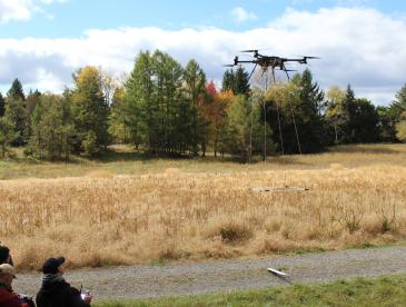 A small team of people operates a drone outdoors, in an area with fields and woods.