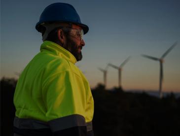 A person wearing a bright yellow high‑visibility jacket, dark safety gloves, and a blue hard hat stands outdoors at dusk, holding a closed laptop at their side. In the distance, several wind turbines rise above a darkened landscape under a fading sky
