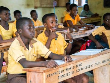 Children sitting at desks from the World Bank. One is smiling and giving a double thumbs up.